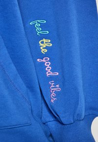 Der blaue Sweatshirt besteht aus weichem Stoff, hat gerippte Bündchen und bestickten Text in mehrfarbigen Fäden mit der Aufschrift "spüre die gute Stimmung".