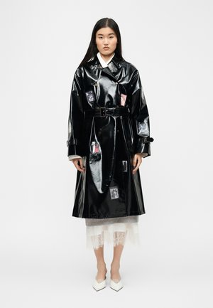 Nő csillogó fekete, öves ballonkabátban, amelyen átlátszó zsebek vannak, fehér csipkeruha és fehér hegyes orrú magas sarkú cipő fölött, egyszerű háttér előtt állva.