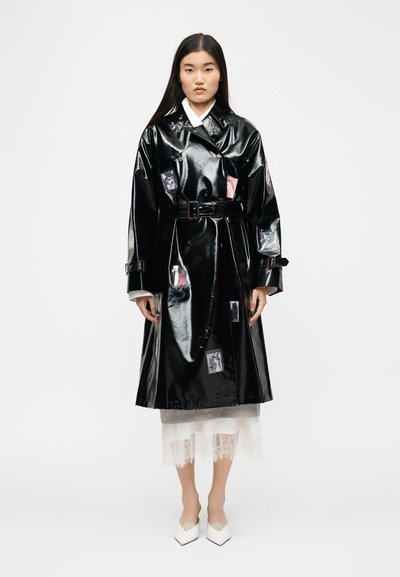 Femme portant un trench-coat noir brillant ceinturé avec des poches transparentes, sur une jupe en dentelle blanche et des talons blancs pointus, debout devant un fond uni.