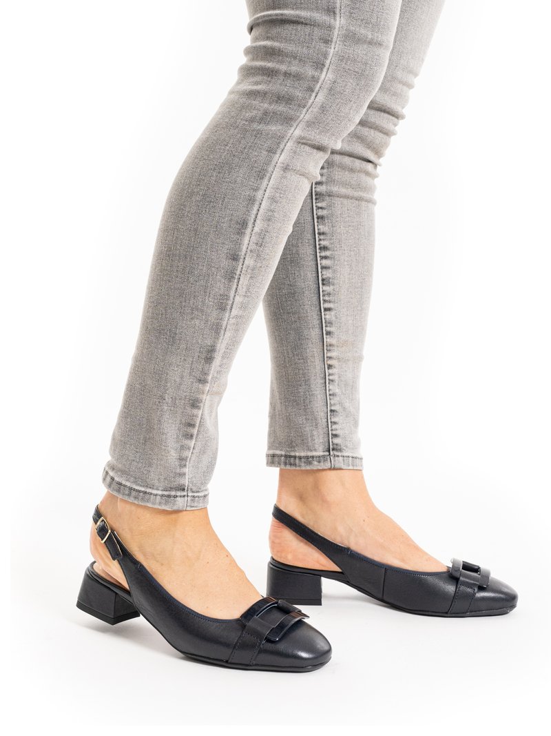 Persona che indossa jeans skinny grigi e scarpe slingback nere con tacco basso e fibbia decorativa, in piedi su uno sfondo bianco.