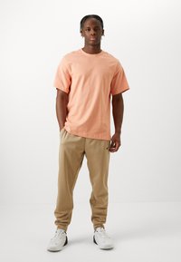 T-shirt en coton corail à manches courtes, associé à un pantalon de survêtement beige avec poches latérales et logo noir, porté avec des baskets blanches.
