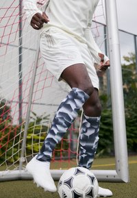 Camiseta y pantalones cortos deportivos blancos con tres rayas; calcetas altas hasta la rodilla con un patrón geométrico en azul, gris y blanco; botas de fútbol blancas; balón de fútbol en blanco y negro.