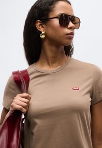 Femme portant un t-shirt marron Levi's, de grandes boucles d'oreilles en or, des lunettes de soleil foncées, tenant une lanière de sac en cuir rouge sur son épaule.