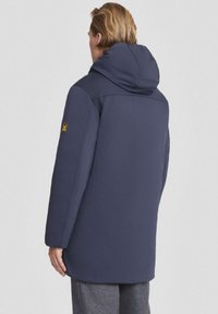 Cappotto isolante blu navy con cappuccio, caratterizzato da una texture liscia, una vestibilità ampia e un logo sulla manica sinistra. Visuale posteriore mostrata.