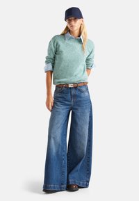 Kobieta nosząca granatową czapkę, jasnoniebieską koszulę pod miętowym swetrem, szerokie niebieskie dżinsy, brązowy pasek i brązowe buty, stojąca z jedną ręką w kieszeni.