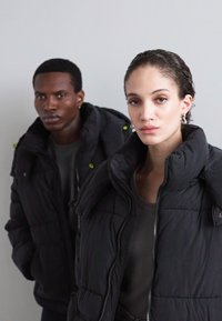 Two individuals wearing black puffer jackets with high collars. Jackets feature quilted texture and green accents. Background is plain.