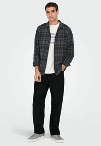 Ung man som bär en mörkgrå rutmönstrad skjorta över en vit grafisk t-shirt, svarta byxor och grå sneakers, står med en hand i fickan.
