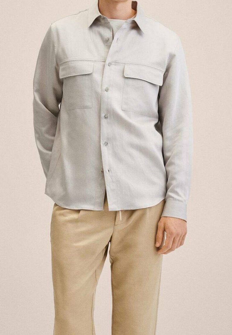 Camisa de botones gris claro en una tela suave, con dos bolsillos en el pecho, cuello clásico y dobladillo recto, combinada con pantalones beige.