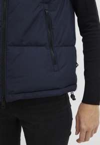 Marineblå vadderad väst med dragkedja framtill, sidofickor och dragsnöre i fållen; slät textur och lättviktig material.