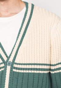 Cárdigan de punto con textura de canalé, bloques de color crema y verde azulado, diseño de escote en V y un botón azul redondo en el frente.