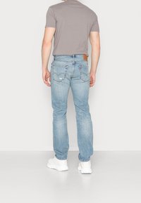 Ljusblå denimjeans med rak passform, fem fickor, lätt slitage och en brun läderlapp på den bakre midjan. Bärs med en grå skjorta.