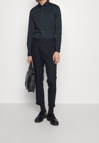 Černá vzorovaná košile s dlouhými rukávy, spárovaná s tmavými kalhotami a černými botami. Doplňky zahrnují černou tašku. Čistý, upravený vzhled.