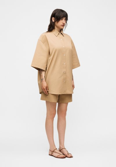 Γυναίκα που φοράει υπερμεγέθη καφέ πουκάμισο με κουμπιά, ταιριαστά σορτς και καφέ σανδάλια με λουριά και επίπεδη σόλα, στέκεται μπροστά σε απλό λευκό φόντο.