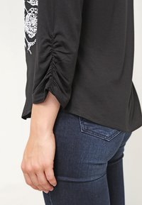 Černý dlouhý rukáv s řasením na rukávu, kombinovaný s tmavě modrými džíny, z pohledu bokem, ukazuje torzo a paži osoby.