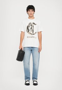 Fehér póló nagy barna és fekete logóval, világoskék farmernadrág és fekete kézi táska. Fekete cipők fehér díszítéssel.