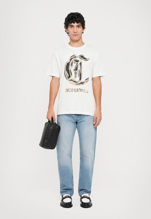 White t-shirt with a large brown and black logo, light blue jeans, and a black handheld bag. Black shoes with white accents.
