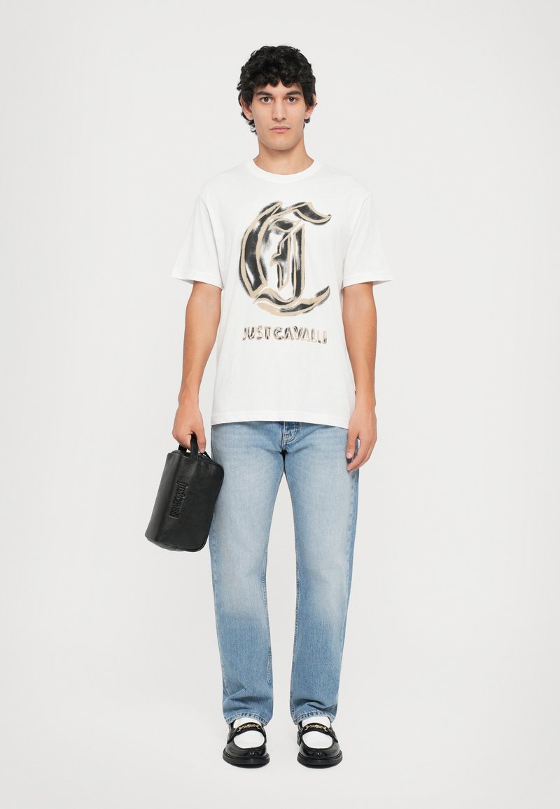 Fehér póló nagy barna és fekete logóval, világoskék farmernadrág és fekete kézi táska. Fekete cipők fehér díszítéssel.