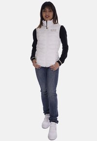 Gilet rembourré blanc avec col haut, fermeture éclair et logo EA7. Porté sur un haut noir à manches longues, associé à un jean bleu et des baskets blanches.