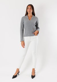 Maglione grigio a coste con scollatura a V, abbinato a pantaloni bianchi aderenti e décolleté nere a punta, su uno sfondo bianco semplice.