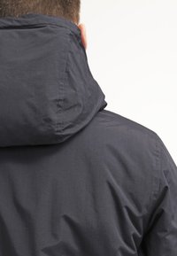 Giacca nera con cappuccio e finitura opaca, caratterizzata da un alto colletto e una texture liscia, vista da dietro.