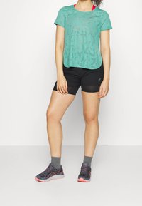 Jasnozielona, siateczkowa bluzka z krótkim rękawem i wyciętymi wzorami, zestawiona z czarnymi szortami. Model ma na sobie szare skarpetki oraz czarne buty sportowe z różowymi akcentami.