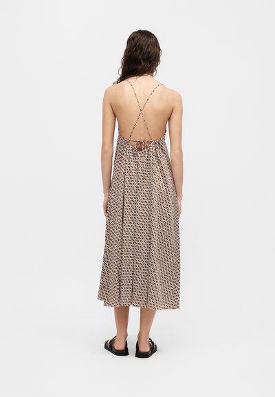Γυναίκα με μακριά μαλλιά που φοράει midi φόρεμα με γεωμετρικό μοτίβο και ανοικτή πλάτη, λεπτές, σταυρωτές τιράντες και μαύρα σανδάλια, στάση σε λευκό φόντο.