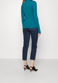 Camisa de manga larga color azul verdoso con corte ajustado, combinada con unos vaqueros oscuros recortados y zapatos de tacón negros. Tejido suave con un ligero brillo.
