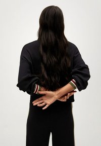 Sweatshirt noir cropped avec des poignets côtelés présentant des rayures vertes, rouges et blanches. Cheveux longs et foncés, mains croisées dans le dos. Pantalons noirs.