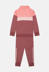 Chándal en tonos rosa y burdeos, que incluye una chaqueta con cremallera y cuello alto, junto con pantalones a juego que tienen franjas laterales y puños acanalados.