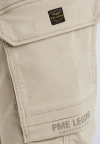 Poche en tissu beige avec rabat sur un vêtement, avec patch de la marque PME Legend et logo imprimé "PME Legend American Classic" près du bord inférieur.