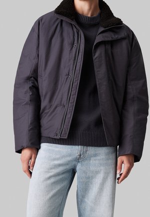 Mężczyzna w ciemnoszarej kurtce rozpinanej na zamek z czarnym kołnierzem z baranka, pod którą ma czarny sweter i jasnoniebieskie dżinsy, na szarym tle.