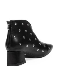 Botín de cuero negro hasta el tobillo con punta afilada, detalles de tachuelas plateadas y cierre de cremallera. Presenta un bajo tacón cuadrado y una textura suave.