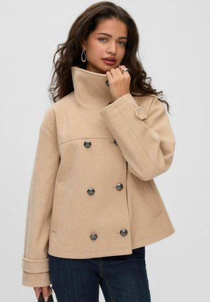 Une femme porte un manteau beige croisé à col montant et à boutons foncés, ainsi qu’un jean, sur un fond uni.