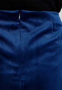 Jupe bleu marine avec une texture lisse et légèrement brillante ; elle présente une fermeture éclair centrée au dos et une silhouette ajustée.