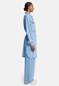 Jasnoniebieska sukienka dżinsowa z długimi rękawami, z przodu na guziki, z dwiema kieszeniami na piersiach i zaokrąglonym dołem. W połączeniu z szerokimi spodniami dżinsowymi w tym samym kolorze i białymi butami.
