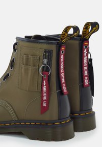 Bottes de combat vert olive et noires avec coutures jaunes, fermeture éclair latérale, et étiquettes rouges "Remove Before Flight" sur la tirette de la fermeture éclair et la boucle du talon.