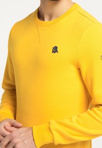 Felpa gialla realizzata in tessuto morbido, con scollo rotondo, polsini a costine e un logo ricamato blu sul petto.