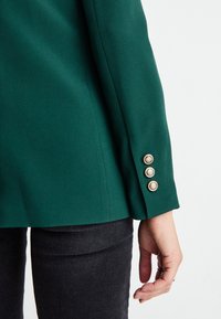 Personne portant un blazer vert foncé avec trois boutons dorés sur la manche et un pantalon gris foncé, main détendue le long du corps.