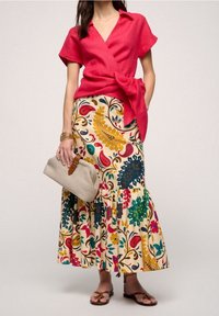 Blusa avvolgente rossa in lino con maniche corte e un fiocco, abbinata a una gonna a strati con motivo floreale in crema, completata da una pochette beige e sandali.