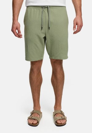 Uomo che indossa pantaloncini verde oliva con coulisse e sandali color cuoio con doppia fibbia, in piedi davanti a uno sfondo bianco.
