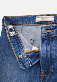 Denim teksad, millel on sinine värv ja kollased õmblused, nööbikinnitus ja vöökohamärk; tekstuuriga kangas koos sisepocket-i üksikasjaga.