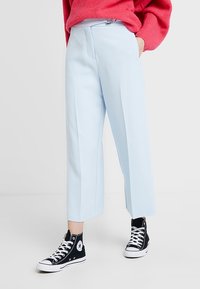 Pantalons cropped bleu clair, ajustés, avec des plis avant et une fermeture éclair, associés à des baskets montantes noires.