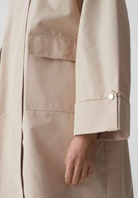 Cappotto beige leggero con maniche lunghe, un singolo bottone al polsino e grandi tasche frontali. Texture liscia e vestibilità comoda.