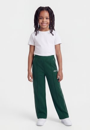 Ung pige med flettet hår iført en hvid t-shirt, mørkegrønne træningsbukser med sideløbende striber og hvide sneakers, stående og smilende.
