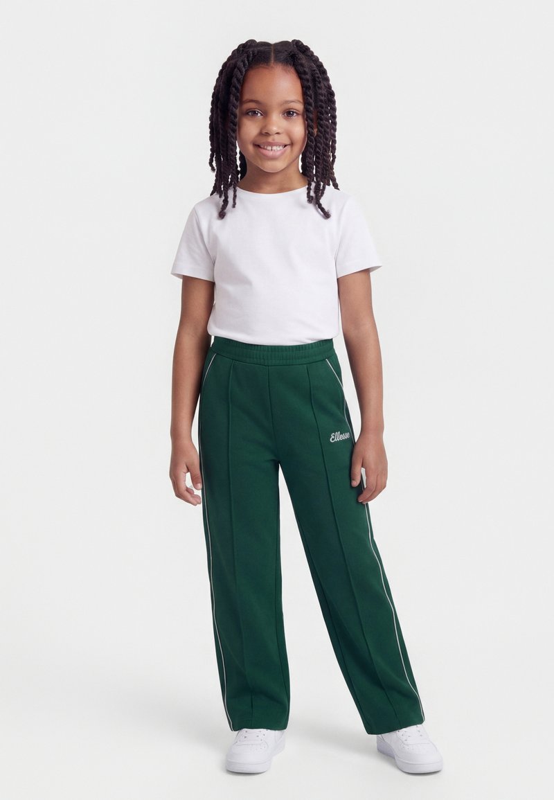 Νεαρή κοπέλα με πλεκτά μαλλιά, φορώντας λευκό μπλουζάκι, σκούρο πράσινο αθλητικό παντελόνι με ρίγες στο πλάι και λευκά αθλητικά παπούτσια, στέκεται και χαμογελάει.