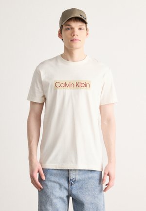 Camiseta de algodón color crema con mangas cortas y un logotipo rectangular de "Calvin Klein" en rojo y beige. Llevada con jeans de mezclilla azul claro.