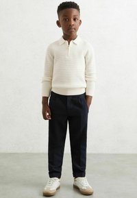 Biele pletené svetrové tričko s krátkym zipsom, námornícke nohavice a béžové tenisky. Svetrové tričko má textúrovaný vzor a rebrované manžety a lem.