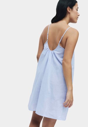 Femme de profil portant une robe ample bleu clair avec des bretelles fines réglables et un dos décolleté, sur un fond clair uni.