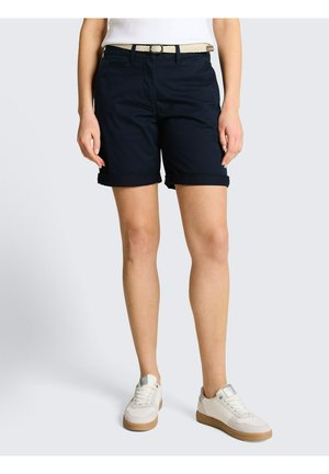 Kobieta ubrana w granatowe, podwinięte szorty z beżowym plecionym paskiem, białą koszulkę i białe trampki z gumowymi podeszwami, stoi na jednolitym tle.