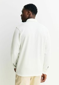 Weißes, geripptes Hemd mit Kragen, lange Ärmel und Knopfmanschetten, von hinten gezeigt. Glatte Textur, lockere Passform.